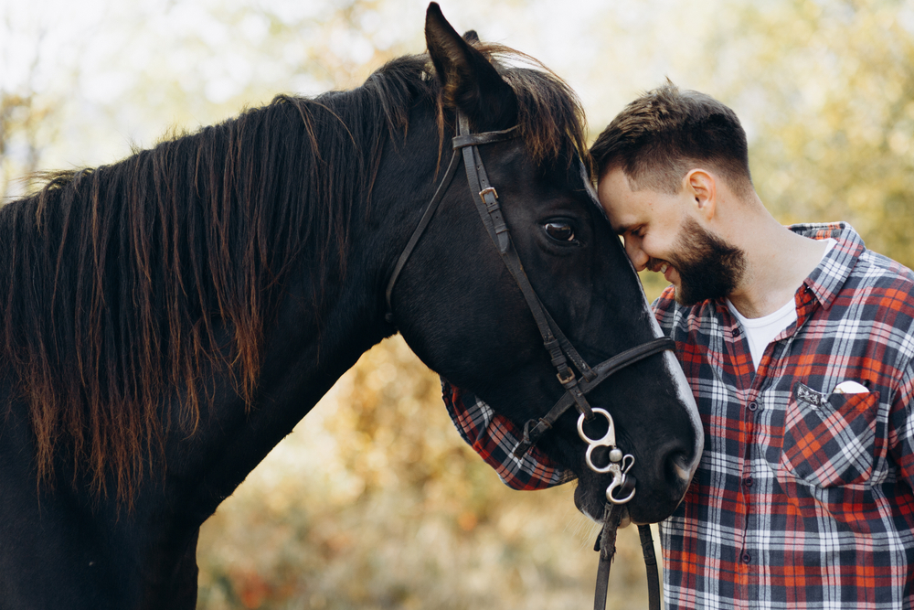A man in a plaid shirt leans his forehead against a black horse, sharing a quiet moment of connection outdoors - Equine Retirement Homes
