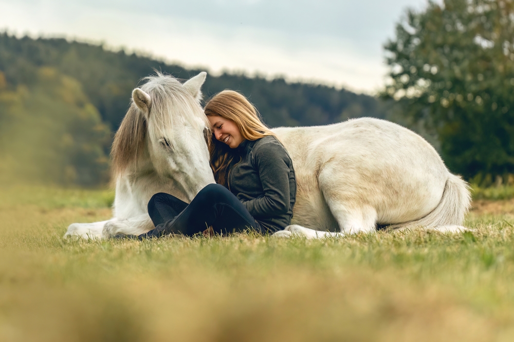 A woman sits in the grass, leaning affectionately against a white horse lying beside her in a peaceful outdoor setting - Equine Retirement Homes
